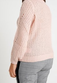 Pull tricoté rose clair avec une maille lâche et ouverte et un ourlet côtelé, présentant des manches larges et une coupe décontractée, associé à un pantalon à carreaux.