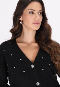 Cárdigan negro de punto con cuello en V, adornado con rhinestones y botones plateados. Textura suave con un diseño de punto texturizado en todo el prenda.