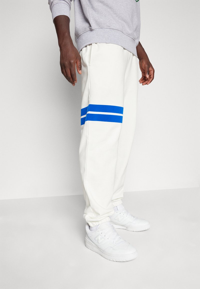 Pantaloni della tuta bianchi con righe blu lungo i fianchi, dotati di una vestibilità comoda e polsini elasticizzati. Abbinati a sneaker bianche.