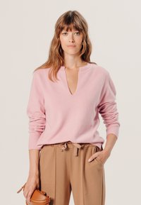 Hellrosa gestrickter Pullover mit V-Ausschnitt, gerippten Ärmeln und lockerer Passform, kombiniert mit beigen Hosen und einer kleinen braunen Handtasche.