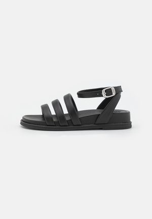 Sort flad sandal med tre frontremme, en polstret sål og en ankelrem med en sølvspænde på hvid baggrund.