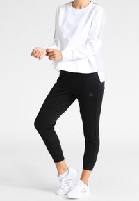 Sudadera blanca de manga larga con aberturas laterales, combinada con pantalones joggers negros que tienen un logo de adidas y zapatillas deportivas blancas.