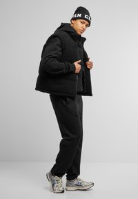 Gilet in pile nero indossato sopra un felpa nera con cappuccio e pantaloni, abbinato a sneakers argento e blu. Il gilet presenta una chiusura con zip e un tessuto testurizzato.