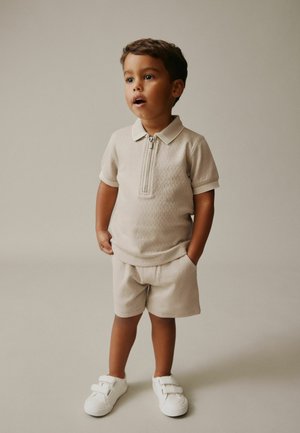 Beige gestricktes Polohemd mit halbem Reißverschluss, dazu passende Shorts, beide mit strukturierten Mustern. Weiße Sneaker mit Riemen vervollständigen das Outfit.
