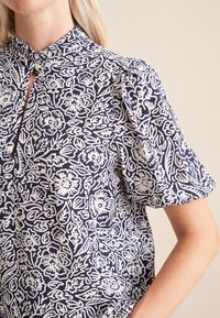 Blusa navy con motivo floreale bianco, caratterizzata da un colletto alto, maniche corte a sbuffo e una apertura a chiave sul collo. Tessuto in cotone.