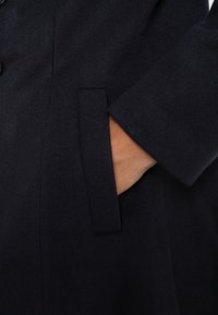 Main partiellement insérée dans la poche d'un manteau en laine foncée, montrant la manche et la texture du tissu.