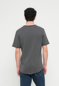 Person with short curly hair wears a plain dark gray short-sleeve shirt and blue jeans, facing a white wall.