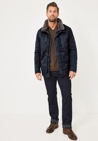 Marineblaue Jacke mit einem Kunstfellkragen, Knopf- und Reißverschlussverschluss, zwei Brusttaschen, kombiniert mit einem braunen Pullover und dunklen Jeans. Braune Stiefel.