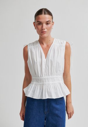 Woman wearing sleeveless white gathered blouse with V-neck and peplum hem, paired with high-waisted blue jeans, standing against plain background.