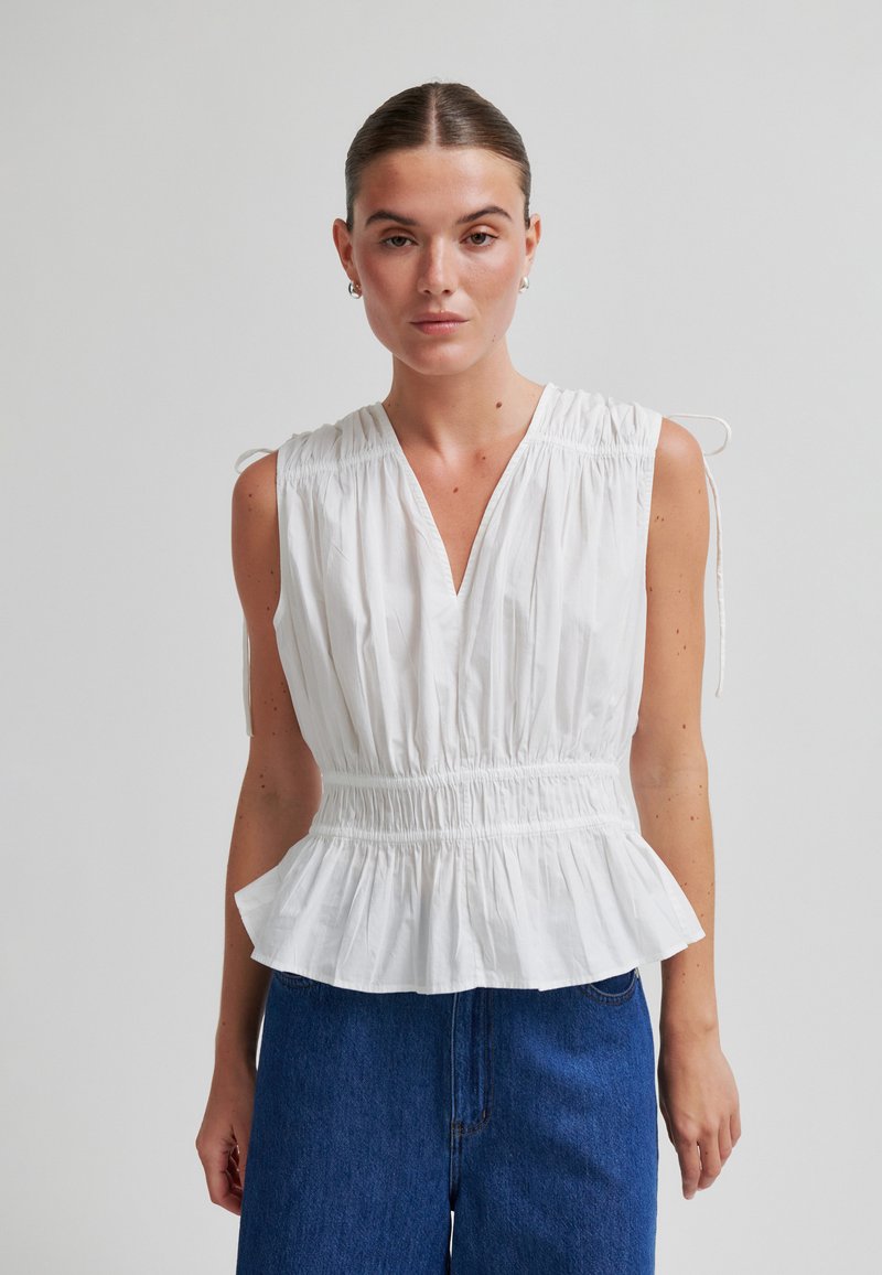 Femme portant un haut blanc sans manches à fronces avec col en V et ourlet péplum, associé à un jean bleu taille haute, debout devant un fond uni.