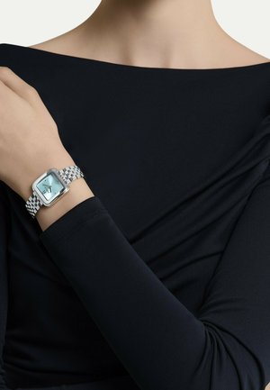 Kvinde iført et sølvfarvet armbåndsur med blå firkantet urskive, klædt i en sort langærmet off-shoulder top.