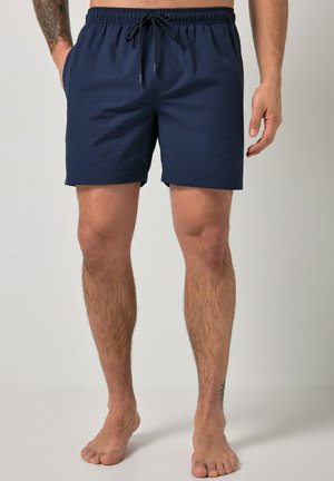 Osoba z gołymi nogami, ubrana w granatowe, teksturowane kąpielówki z elastycznym pasem i sznurkiem, stojąca na jednolitym tle.