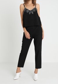 Haut cropped noir avec broderie florale argentée, associé à un pantalon noir taille haute. Le modèle porte des mules blanches à talons.
