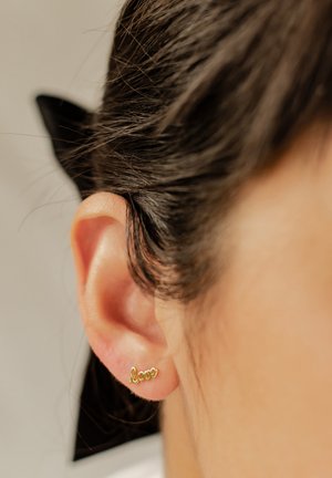 Gold earrings in a cursive "love" design, worn on the lobe. The ear has smooth skin and dark hair pulled back.