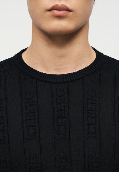 Bas du visage et cou d'une personne portant un pull noir côtelé avec le texte "ICEBERG" tricoté verticalement sur le tissu.