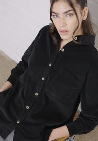 Zwarte corduroy blouse met knopen en een borstzak, voorzien van een puntige kraag en contrasterende knopen. Zachte textuur met een relaxte pasvorm.