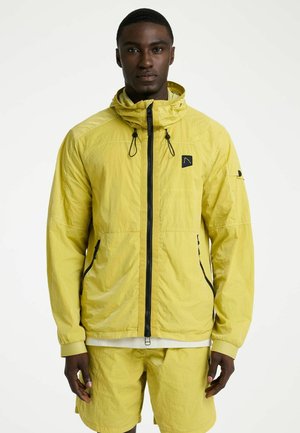 Man draagt een geel capuchonjack met zwarte rits en bijpassende gele korte broek, staand tegen een effen achtergrond.