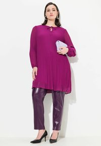 Plissé paarse tuniek met sleuteldetail, gecombineerd met glanzende bordeauxrode pantalons en zwarte hoge hakken, met een lichtpaarse clutch.