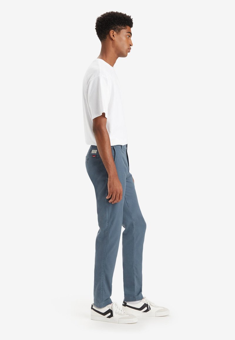 Pantalons en coton stretch bleu avec une coupe slim, présentant une jambe droite, des poches latérales et un patch de marque sur la poche arrière. Portés avec un t-shirt blanc.