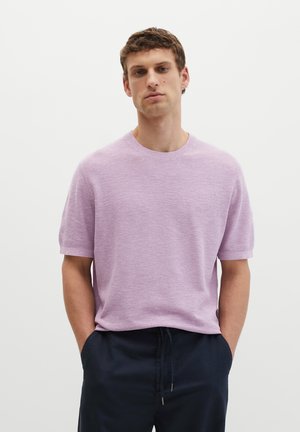 Jonge man staand met handen in de zakken, draagt een lichtpaarse korte mouw trui en donkerblauwe broek voor een effen witte achtergrond.