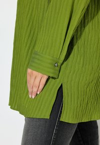 Maglia verde a coste a maniche lunghe con polsini abbottonati, caratterizzata da fessure laterali e tessuto testurizzato, abbinata a jeans scuri.