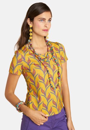 Woman in a yellow patterned shirt with purple pants, wearing long colorful beaded necklaces, yellow floral earrings, and multicolored bracelets.