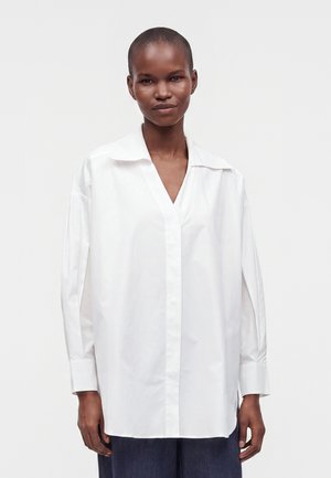 Persona che indossa una camicia bianca a maniche lunghe con colletto e pantaloni scuri, in piedi davanti a uno sfondo chiaro e uniforme.