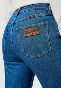 Modri kavbojki z teksturiranim zaključkom, ki imata rjav usnjen našitek z logotipom "Wrangler" in značilnim rumenim šivom na zadnjih žepih.