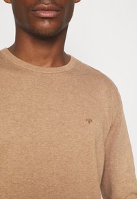 Mężczyzna w jasnobrązowym swetrze typu crew neck z małym haftowanym logo na piersi, na jednolitym tle.