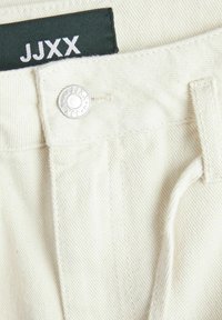 Tejido de mezclilla blanca con textura visible, que presenta un botón plateado y una etiqueta verde con "JJXX" en letras blancas.