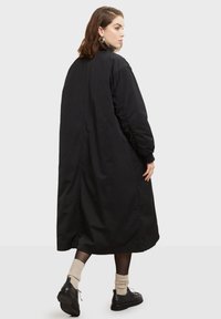 Lang sort frakke med en afslappet pasform, lavet af blødt stof. Har rynkede ærmer og længde til midt på læggen, med sideslids for let bevægelse.