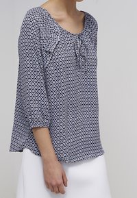 Blouse bleue et blanche à motifs avec un col rond, des manches trois-quarts et un détail à nouer à l'avant, fabriquée en tissu léger.