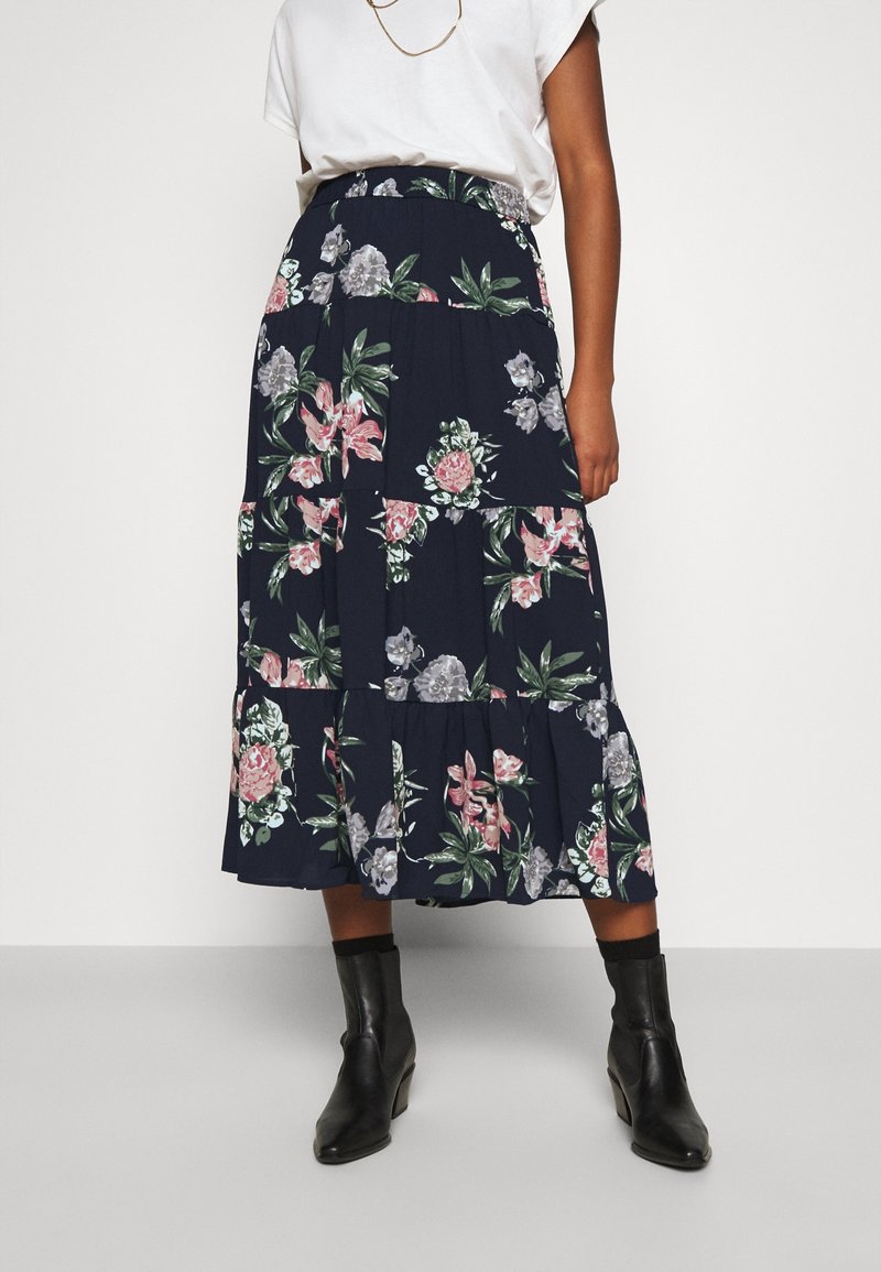 Femme portant une jupe midi marine à fleurs roses et grises, un t-shirt blanc rentré, des bottines noires et un collier en or.