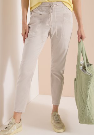 Osoba nosząca jasnobeżowe spodnie ze sznurkiem, beżowe trampki, żółtą bluzkę, trzymająca dużą pikowaną zieloną torbę przy boku.