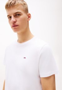 Młody mężczyzna o krótkich blond włosach, ubrany w prostą białą koszulkę z małym logo w kolorach czerwonym, białym i niebieskim na piersi.