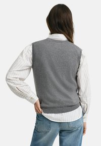 Un gilet senza maniche grigio indossato su una camicia a righe bianche e rosse, abbinato a jeans blu. Tessuto liscio, vestibilità classica e orlo a coste.