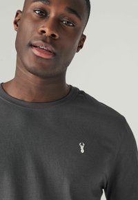 Grå T-shirt lavet af blødt stof, med rund hals og et lille broderet logo på venstre bryst. Glat overflade og minimalistisk design.