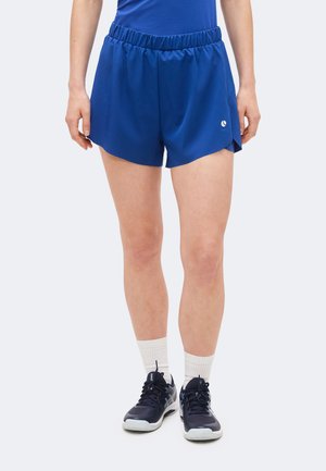 Persoon die blauwe sportshorts met elastische taille, witte sportsokken en zwarte sportschoenen draagt, staand tegen een lichte achtergrond.