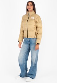 Beige pufferjack met hoge kraag, rits aan de voorkant en logo op de borst. Gekenmerkt met lichtblauwe wijde jeans en witte sneakers.