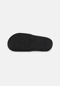 Black rubber sandal sole with a textured pattern, featuring the embossed "LACOSTE" logo. Rounded shape with a non-slip tread design.