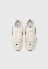 Jasno beżowe buty sportowe z siateczkową i syntetyczną cholewką, zaokrąglony nos, wiązany fason oraz czarny logotyp Nike w formie „swoosh” po bokach.