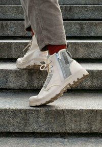 Bottes montantes beiges à finition texturée, avec une fermeture éclair sur le côté, semelle en caoutchouc et panneaux gris contrastés, portées avec des chaussettes rouges.