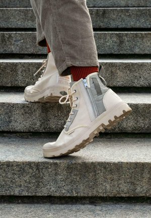 Bottes montantes beiges à finition texturée, avec une fermeture éclair sur le côté, semelle en caoutchouc et panneaux gris contrastés, portées avec des chaussettes rouges.