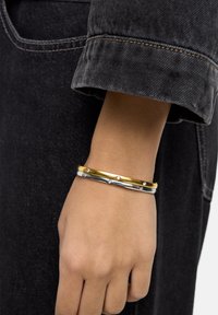 Trzy nałożone bransoletki wykonane ze złota, srebra i bia�łego metalu z małymi diamentowymi akcentami. Prezentowane na nadgarstku na tle ciemnej tkaniny.