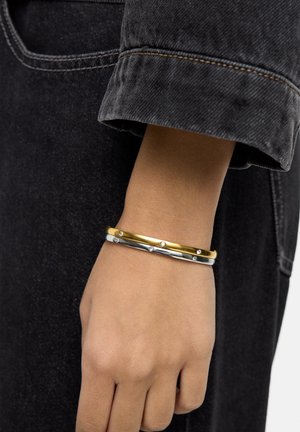 Trzy nałożone bransoletki wykonane ze złota, srebra i białego metalu z małymi diamentowymi akcentami. Prezentowane na nadgarstku na tle ciemnej tkaniny.