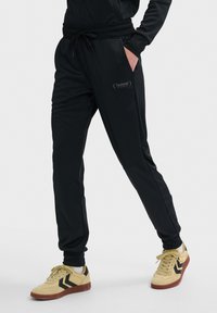 Zwarte sportbroeken met een trekkoord in de taille, zijzakken en een tonale logo op de bovenbeen. Draagt met gele sneaker met zwarte accenten.