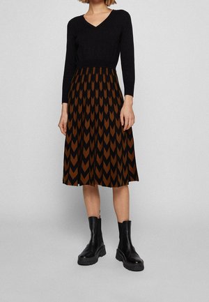 Femme portant un haut noir à col en V à manches longues, une jupe mi-longue à motif chevron marron et noir, et des bottines noires épaisses.