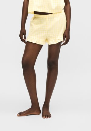 Parte inferior del cuerpo de una persona que lleva pantalones cortos de cuadros amarillos claros con dobladillos con volantes, de pie descalza sobre un fondo blanco liso.