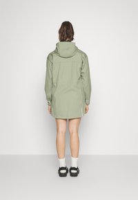 Vrouw die met de rug naar de camera staat, gekleed in een lichtgroene hoodie, witte sokken en zwarte sandalen, tegen een effen witte achtergrond.