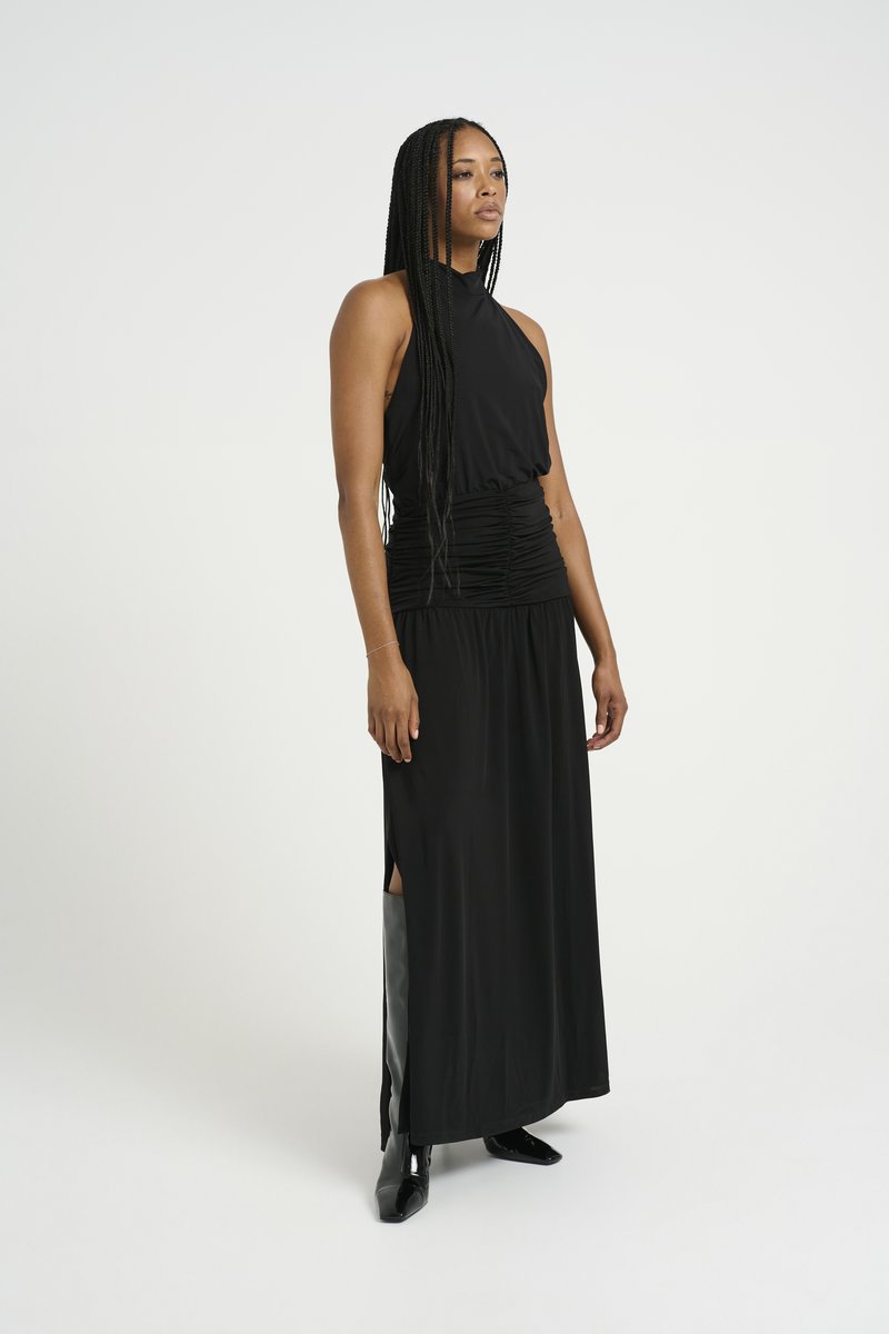 Μαύρο φόρεμα με λαιμόκοψη halter και ελαστική μέση, ρευτούς υφάσματος, πλαϊνές σχισμές και μαύρα υποδήματα μέχρι τον αστράγαλο. Λείος υφής και μίνιμαλ σχέδιο.